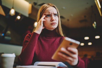 Young beautiful pensive talented female student feeling exhausted while preparing for examinations with modern device indoors.Charming blonde woman tired while working in coffee shop using gadget