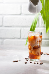 Cold brewed iced coffee in a tall glass cup and coffee beans on a gray concrete background. Pouring milk into a glass of coffee.