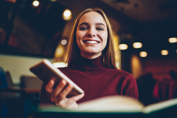Half length portrait of attractive cheerful female blogger enjoying recreation in comfortable coffee shop with modern device.Cute emotional hipster girl sincerely laughing while looking at camera