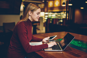 Fototapeta premium Young gorgeous positive intelligent female student looking for information in network while preparing for upcoming exams.Charming happy freelancer reading news in website during remote work indoors