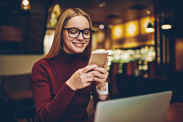 Positive woman in spectacles for provide eyes protection reading text message with good news from colleague, happy successful hipster girl enjoying telephone communication in social networks
