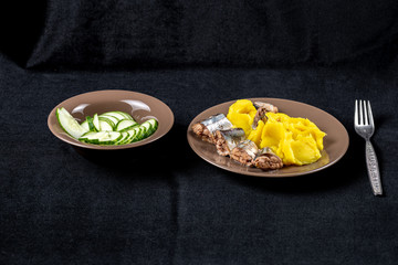 In a deep small plate sliced cucumber in a large flat plate mashed potatoes with saury slices in butter sauce on a table covered with a black cloth with a fork