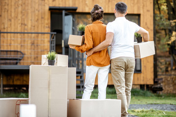 Young couple standing back with cardboard boxes in front of their new house. Concept of a relocation into a new home