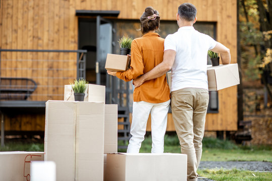 Young couple standing back with cardboard boxes in front of their new house. Concept of a relocation into a new home