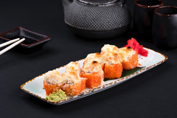Close up of assorted baked sushi set with salmon, shrimp, caviar, mussels, tuna, cream cheese and hot sauce and chopsticks, soy on black background. Hot spicy rolls and green tea teapot
