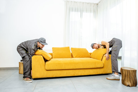 Professional movers in uniform placing yellow sofa at the living room of a new apartment