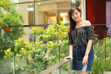 Portrait of Asian beautiful girl wears black off shoulder top and jeans skirt standing by the glass balcony with garden in building. Asian girl model outdoor.