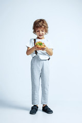 Pretty young curly boy in casual clothes on white studio background. Eating burger. Caucasian male preschooler with bright facial emotions. Childhood, expression, having fun, fast food. Astonished.