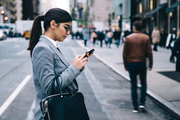Side view of intelligent female employee using cellphone gadget for online chatting during break at urban setting, businesswoman checking account balance on financial web page or application
