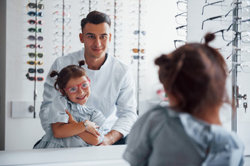 Young pediatrician in white coat helps to get new glasses for little girl