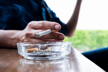 Man keep cigarette in hand, cigarette ends and ash in filthy ashtray on wooden table