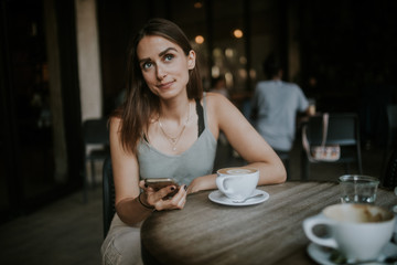 Young woman in thinking pose sitting in a cafe with phone and coffee
