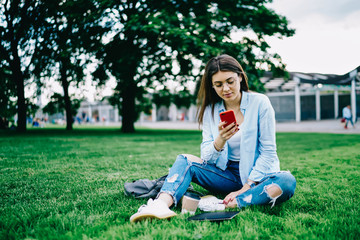 Teen hipster girl checking mail and publicating new post in networks via smartphone on free time in park, young woman recreating on green grass using mobile phone for chatting and reading news