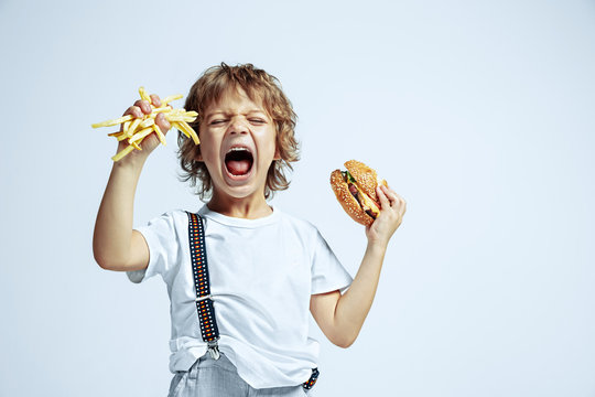 Pretty young curly boy in casual clothes on white studio background. Eating burger with fried potato. Caucasian male preschooler with bright facial emotions. Childhood, expression, fun, fast food.