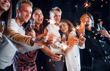 Having fun with sparklers. Confetti is in the air. Group of cheerful friends celebrating new year indoors with drinks in hands