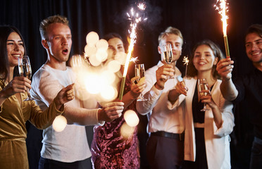 Fototapeta na wymiar Having fun with sparklers. Group of cheerful friends celebrating new year indoors with drinks in hands
