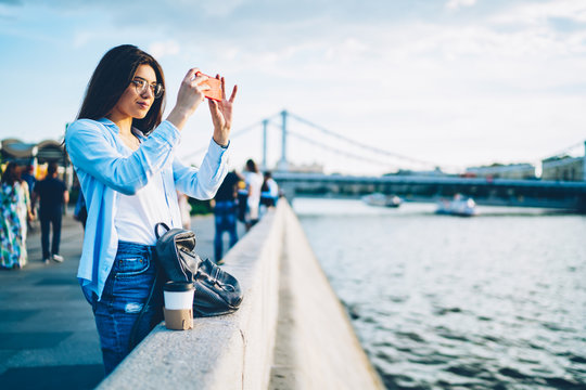 Female tourist in eyewear making photos on modern telephone enjoying beauty of city. Hipster girl taking pictures on river on mobile phone standing on bridge near publicity area for your advertising