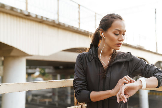 Image of woman using earphones and smartwatch while leaning on railing