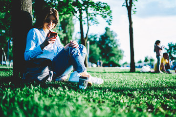 Fototapeta premium Positive hipster girl in trendy casual wear reading message from friend on mobile sitting in park enjoying spring weather, young woman installing application for downloading music from networks