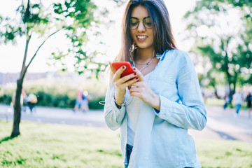 Cheerful hipster girl in eyewear downloading song to smartphone playlist while spending spring day in park, smiling young woman connecting to radio broadcast using mobile phone and earphones on leisur