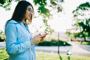 Thoughtful brunette woman in spectacles looking away listening audio book while spending time in park on leisure, hipster girl enjoying music from playlist using cellular and modern earphones