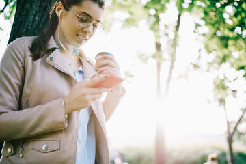 Obraz premium Attractive hipster girl in eyewear checking notification on smartphone download new song to playlist, female meloman listening to music in earphones via app while having coffee break outdoors