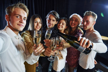 Takes selfie. Group of cheerful friends celebrating new year indoors with drinks in hands
