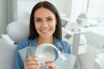 smiling woman sitting in dental chair holding mirror after dental procedure and looking at the camera