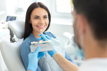 Male dentist holding teeth color palette while beautiful smiling woman looking at him in clinic