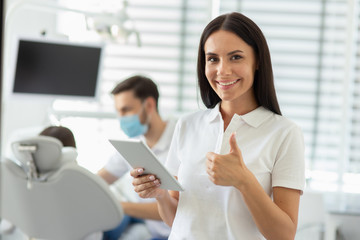 caucasian female doctor showing thumb up, holding digital tablet and smiling at the camera with colleague working with client on the background