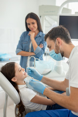 Obraz premium male doctor with instruments in the hands looking girls teeth sitting in the dental chair while mother standing near her for support