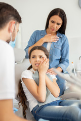 girl with toothache sitting in dental chair while mother standing near her for support