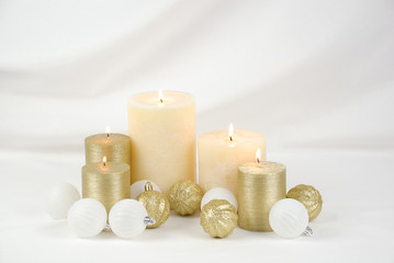 Burning cream and gold Christmas candles with gold and white ball ornaments on a draped white...