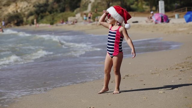 pretty girl toddler wearing Santa hat and trendy sunglasses walks along beach wet sand near rolling warm waves slow motion
