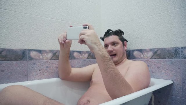 having fun at home.modern weird emotional guy lying in bath uses smartphone