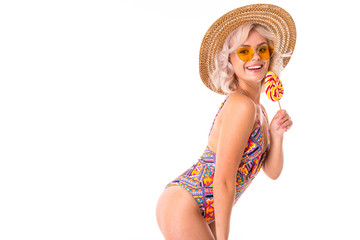 Pretty blonde caucasian female stands in swimsuit with lolipop and smiles isolated on white background