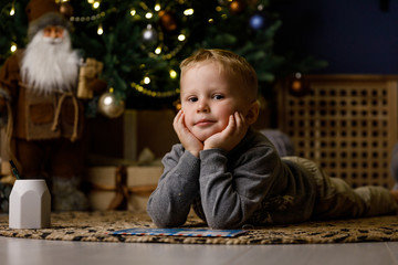 А little cute boy writes a letter to Santa Claus, dresses up a Christmas tree and dreams of a miracle on the background of the Christmas interior.