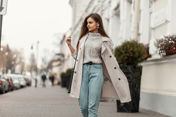 Model of a young beautiful woman in a fashion trench coat in a stylish knitted sweater with a leather black bag walks on the city near a white building. Elegant pretty trendy girl travels the street.