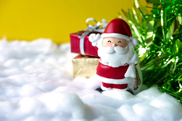 Merry Christmas and Happy New Year concept.Santa Claus doll waving his hand and smile near gift boxes and Christmas tree on snow background.Yellow backdrop and copy space for text.