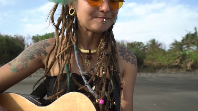 Young hippie girl with girl with colored hair, dreadlocks, piercing and tattoos plays guitar and sings on a beach at sunny day.