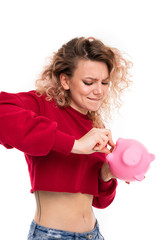 Caucasian girl with curly fair hair trying to get money from the pink pig moneybox, portrait isolated on white background