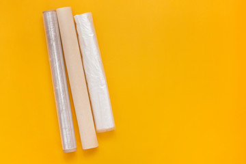 Plastic wrap, transparent package and roll of parchment paper on yellow background. Concept of Packing products in kitchen