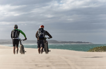 Two cyclists pushing bicycles across sand dunes on the beach