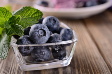 Blueberries in a transparent glass bowl with a sprig of mint on a wooden background near a piece of blueberry cake, close up