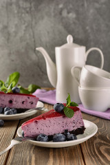 Slices of blueberry mousse cake with mirror glaze decorated berries and leaves of mint next to white teapot and cups, vertical orientation