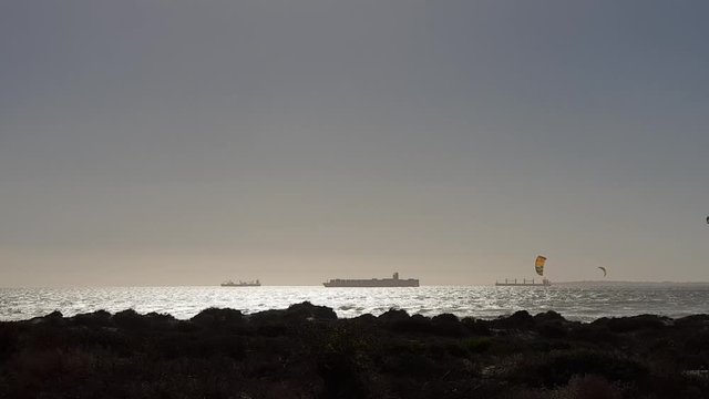 Kites of Cape Town kitesurfers flying behind beach vegetation. Boat silhouette on horizon. Robben Island  visible on right.  Sun glistening on silver ocean waves. Sunlight on mirror sea. Vacation