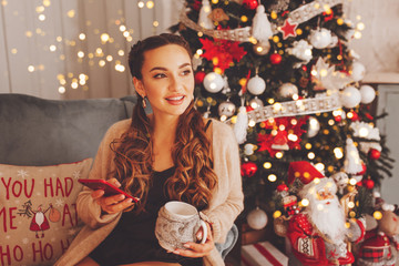 Happy woman sitting on the sofa and looking at window and hold mobile phone. Christmas tree with decoration is next to her. Holidays and technology concepts.