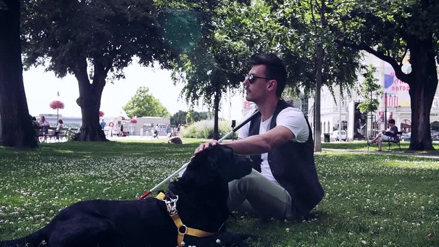 Young blind man with white cane and guide dog sitting in park in city.