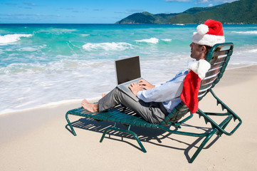 Businessman dressed in Santa hat sitting typing on his laptop on a tropical Caribbean beach