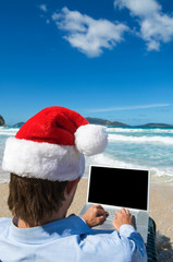 Businessman taking a holiday break in Santa hat as he works on his laptop computer on a tropical beach